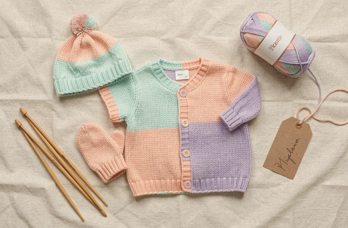 Assortiment de tricots naissance en laine Bebecolor pastel – bonnet, moufles et gilet – posés à plat sur lin crème avec aiguilles à tricoter